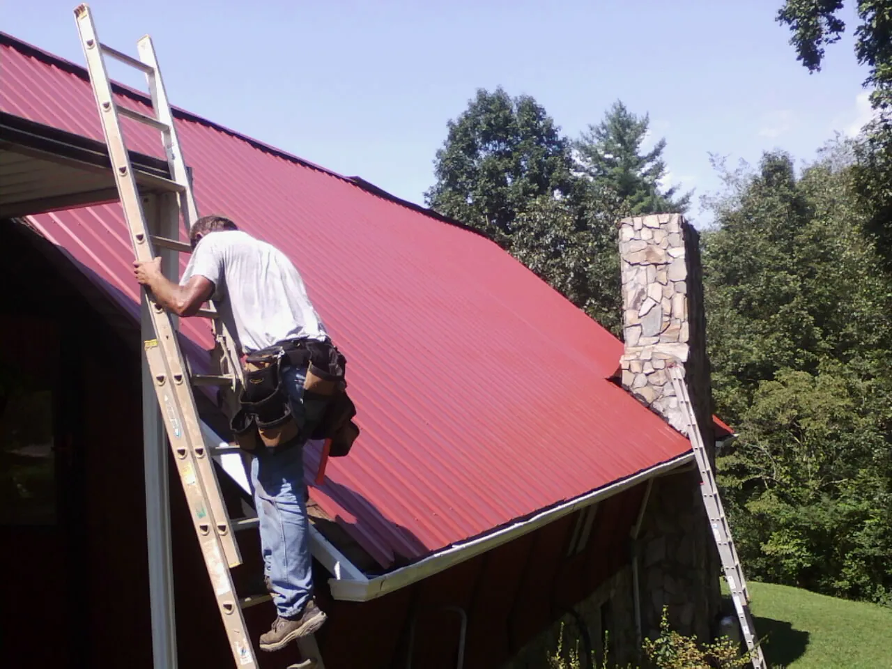 Roofer on a ladder beside a red metal roof and stone chimney — Soffit & Fascia Repair by Roofing Brothers in Burr
